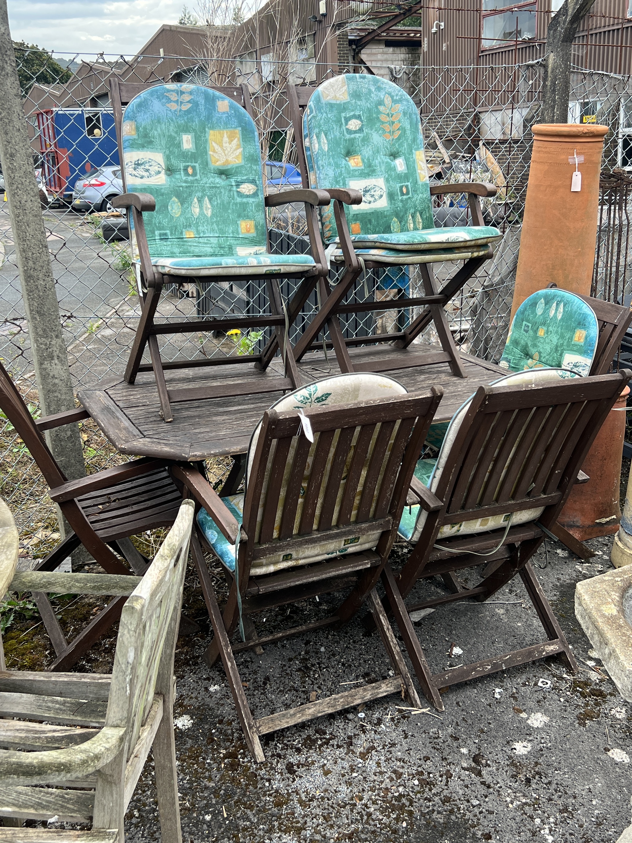 An octagonal stained teak folding garden table, length 150cm, depth 85cm, height 71cm and six folding elbow chairs and five seat pads